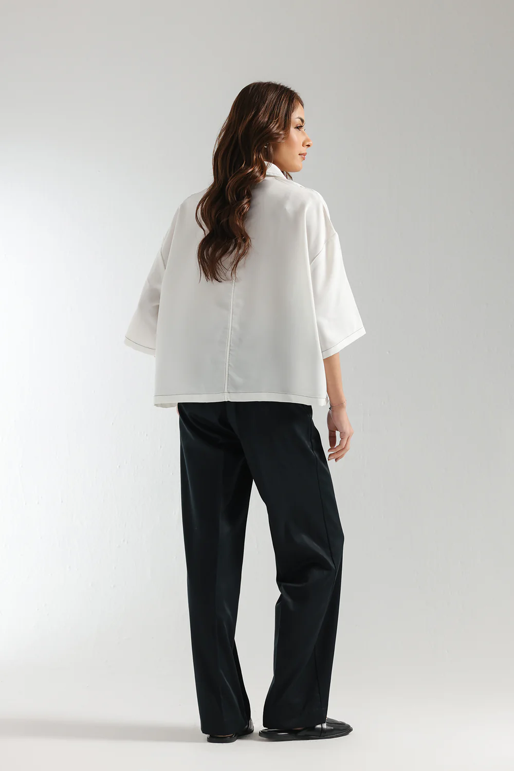 Back studio portrait of Biljiki crisp white boxy button-down shirt with black contrast stitching on a 5'6 model.